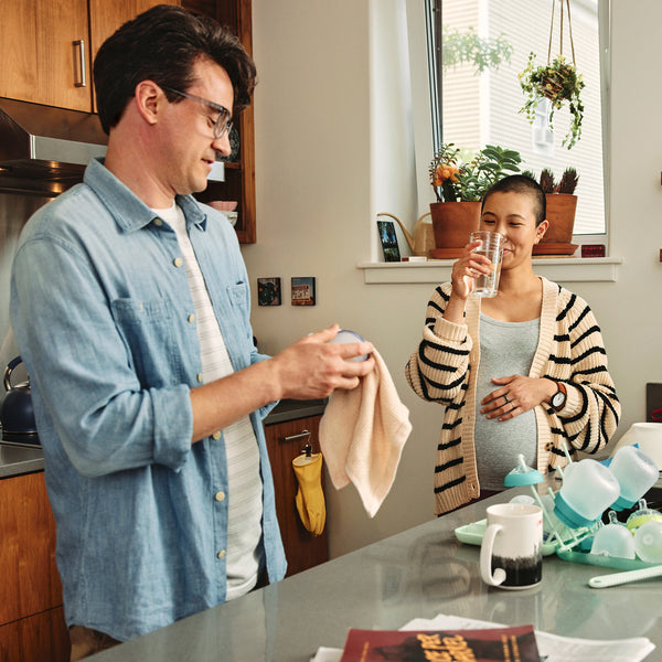 A photo of two smiling individuals in their kitchen. One happily drinks tap water from a glass, while the other cleans their empty cup.