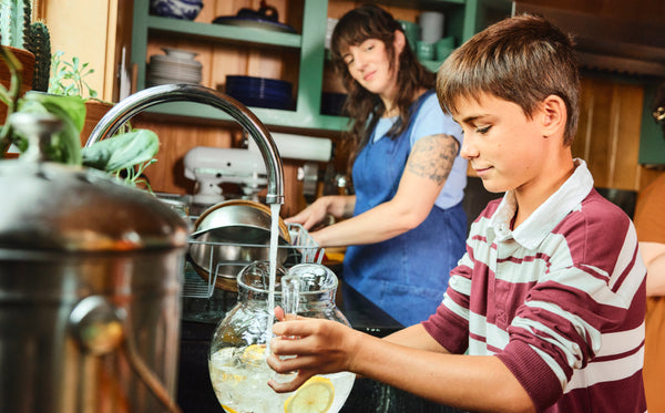 A mother watching her son, a proud look on her face. The son is filling a pitcher of lemonade with fresh, filtered tap water.