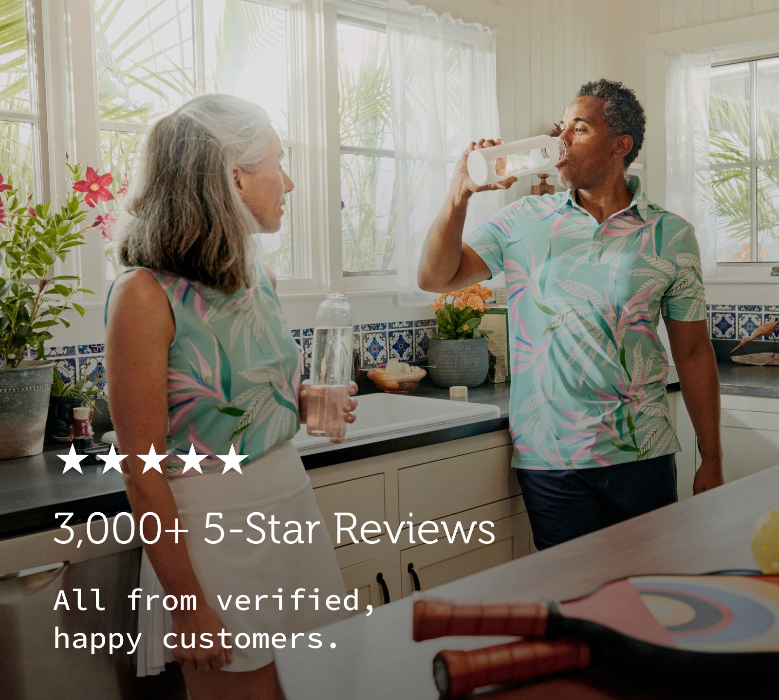 A photo of a couple standing at the kitchen sink. They're filling and drinking their water bottles after a game of pickleball.